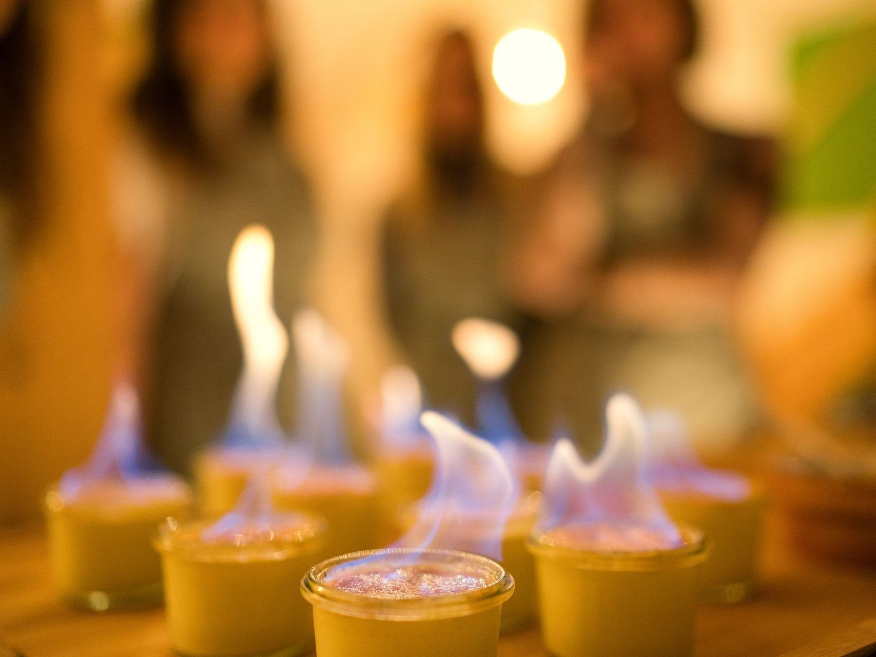 Flambierte Desserts in kleinen Glasgefäßen mit blauen Flammen. Im Hintergrund sind Menschen verschwommen, drinnen.