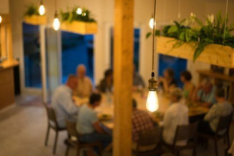 Menschen sitzen bei einem Abendessen um einen Tisch herum, beleuchtet von hängenden Glühbirnen und Blumentöpfen an der Decke.