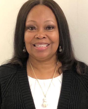 Woman in navy dress, silver necklace, hands clasped, in front of modern metal sculpture.