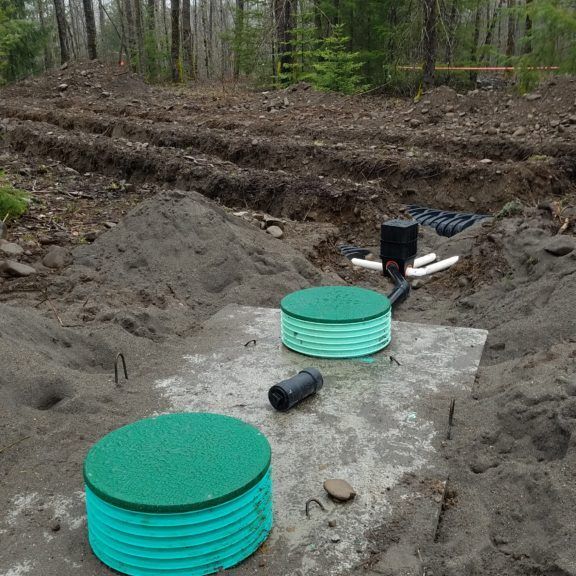 Septic tank installation in dirt, two green-lidded tanks, PVC pipes, and buried trenches in a wooded area.