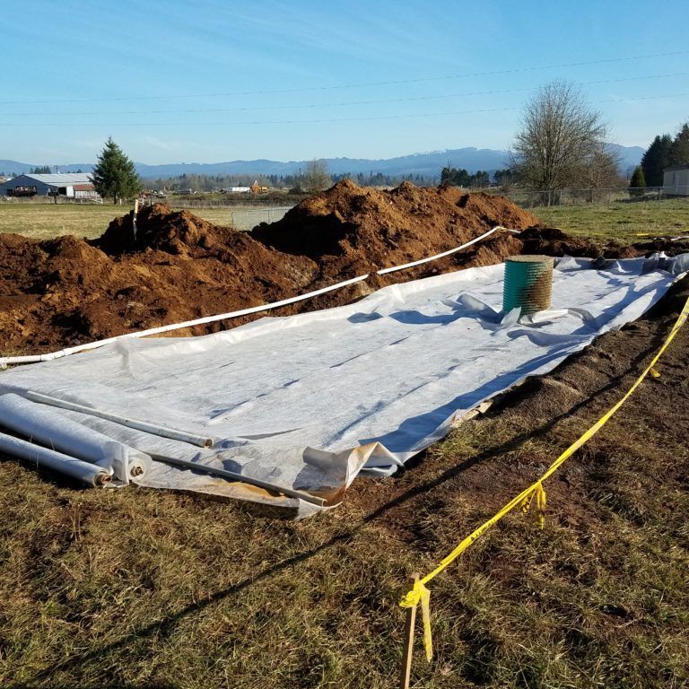 Construction site with white fabric, pipes, a dirt pile, and a green pipe. A field in the background.