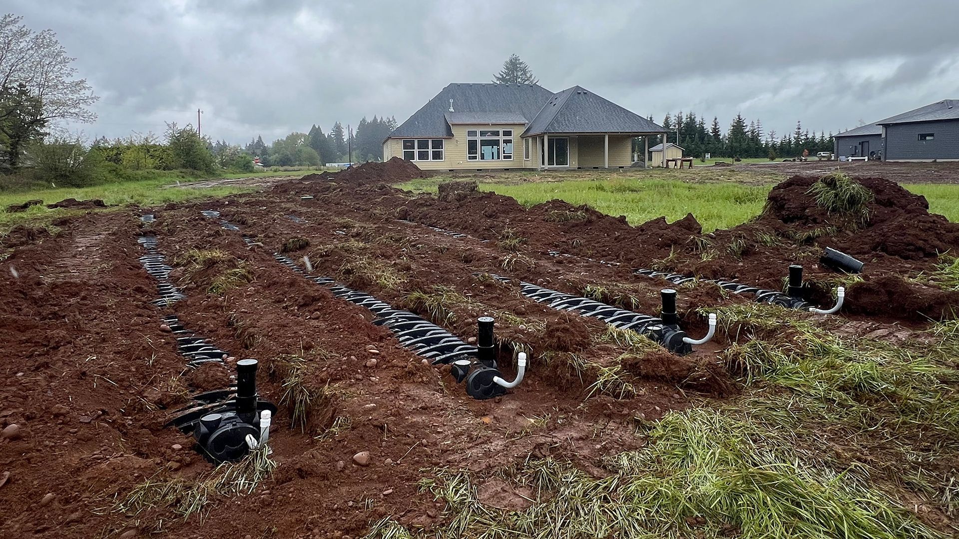Rows of black pipes emerging from freshly dug earth, in front of a house under construction.