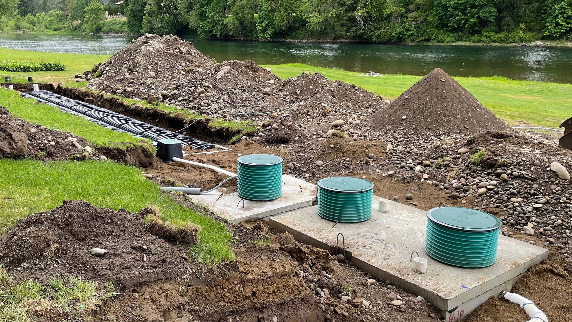 Construction site with septic tanks, pipes, and mounds of dirt near a body of water. Green tanks and grass visible.