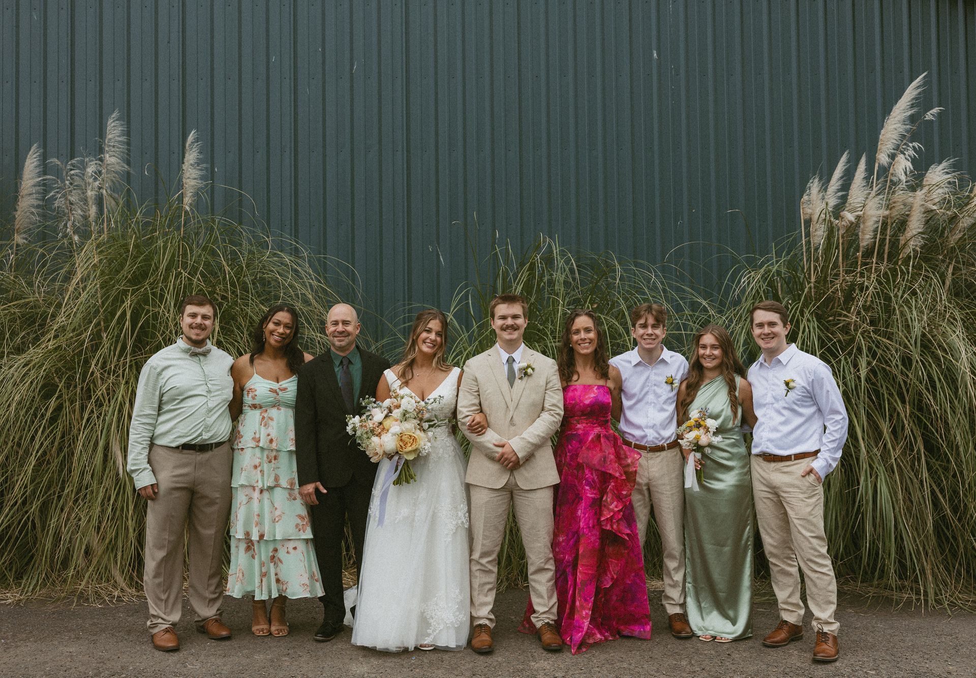 Wedding group: Bride and groom flanked by family, against a teal wall and tall grass.