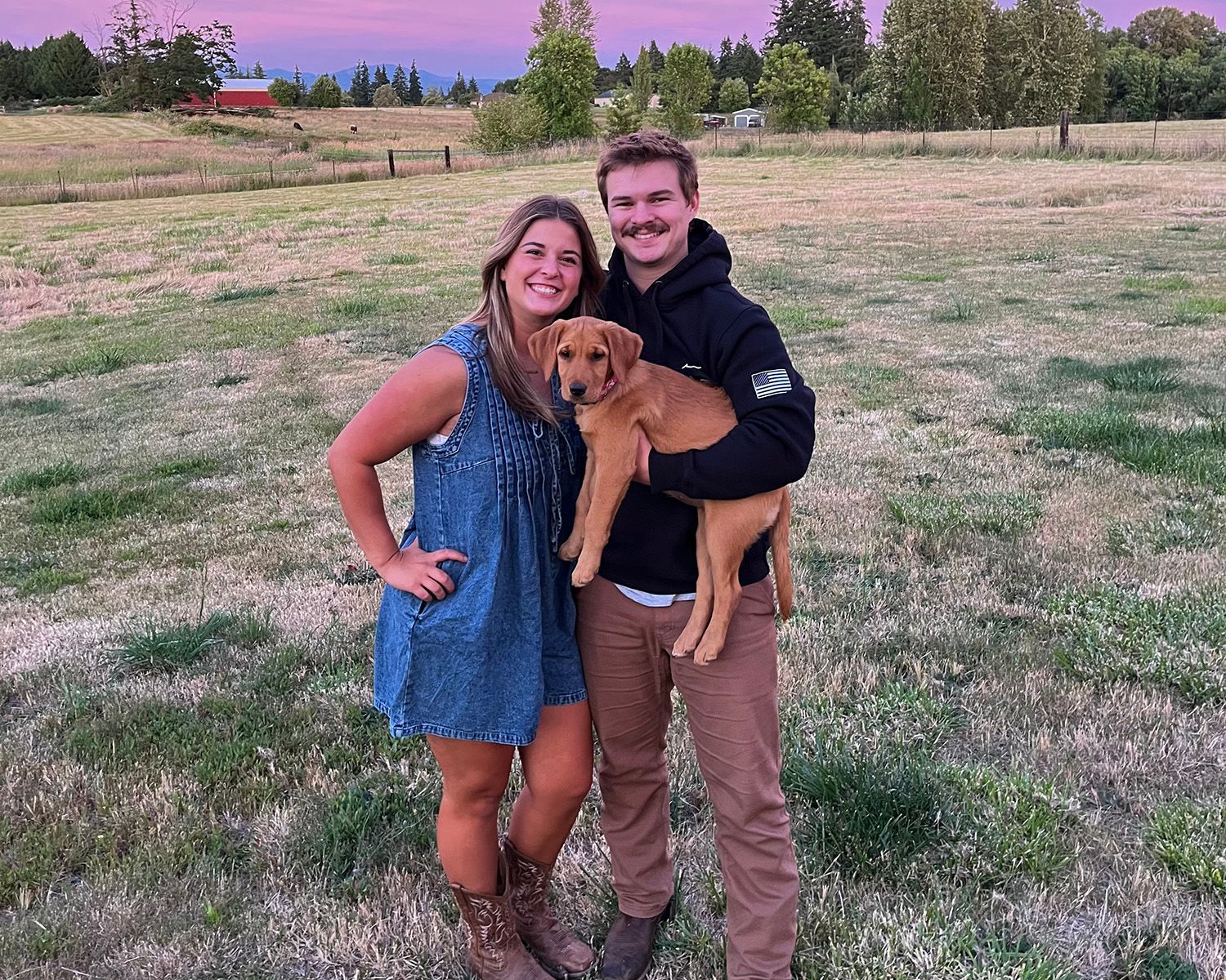 Couple holding a puppy in a field at sunset. Woman in a blue dress, man in a hoodie, and puppy.