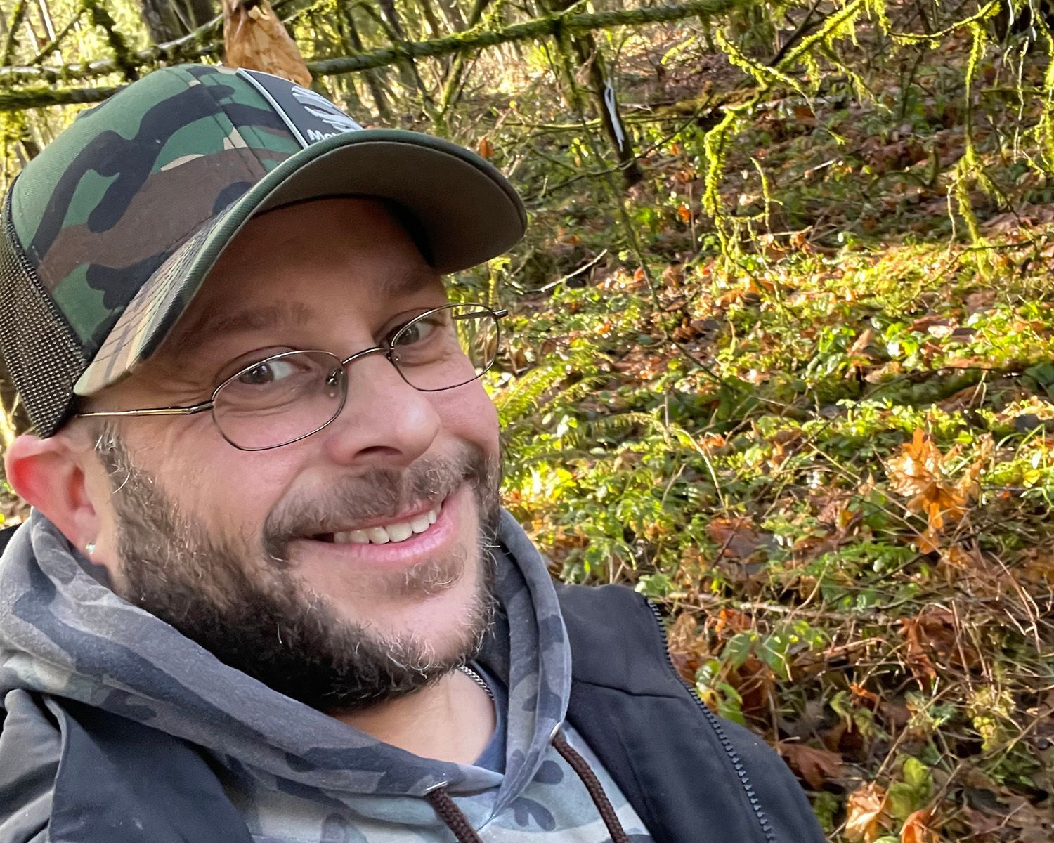 Man with glasses and camo hat smiles outside in a forest.
