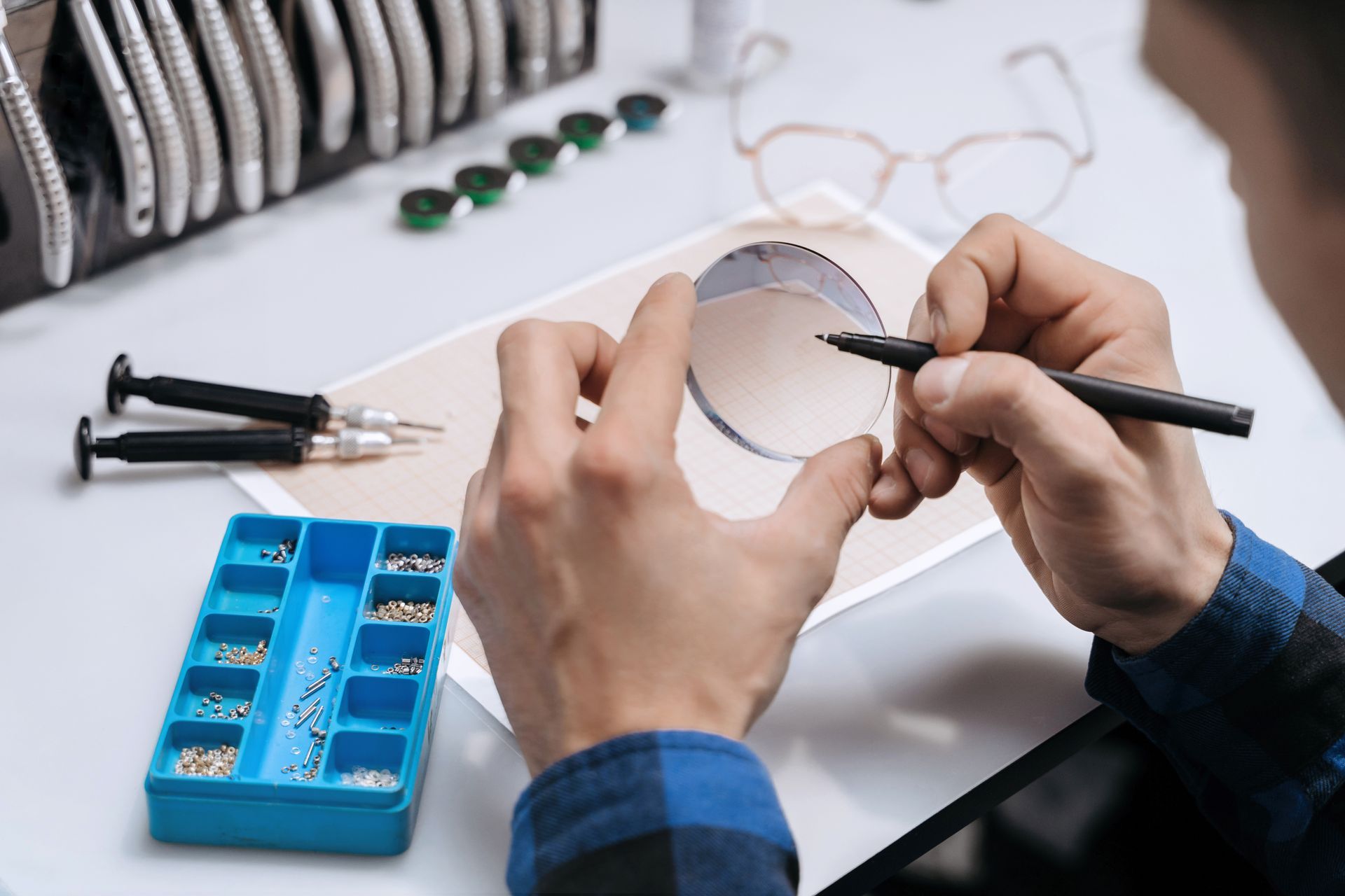 Hands using tweezers to inspect a small round lens at a workbench with tools and a blue organizer.