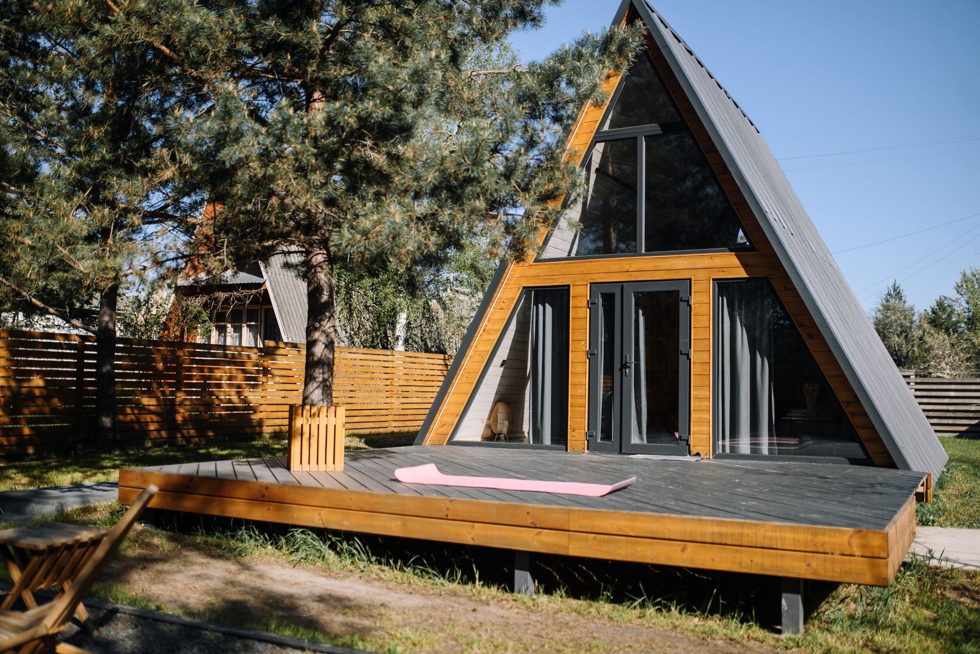 A-frame cabin with a wooden deck. Tall windows and doors. Set in a grassy area, surrounded by trees.