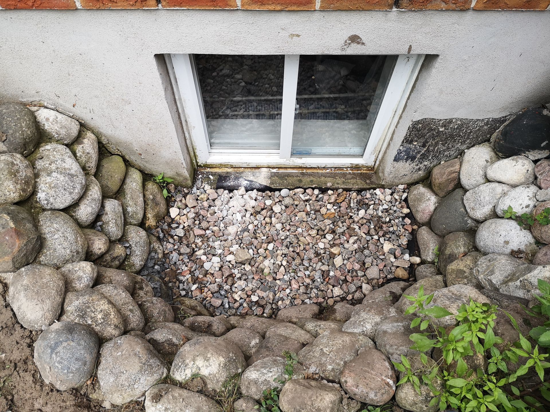 Basement window well with rocks and gravel. Gray stones form the surrounding walls.