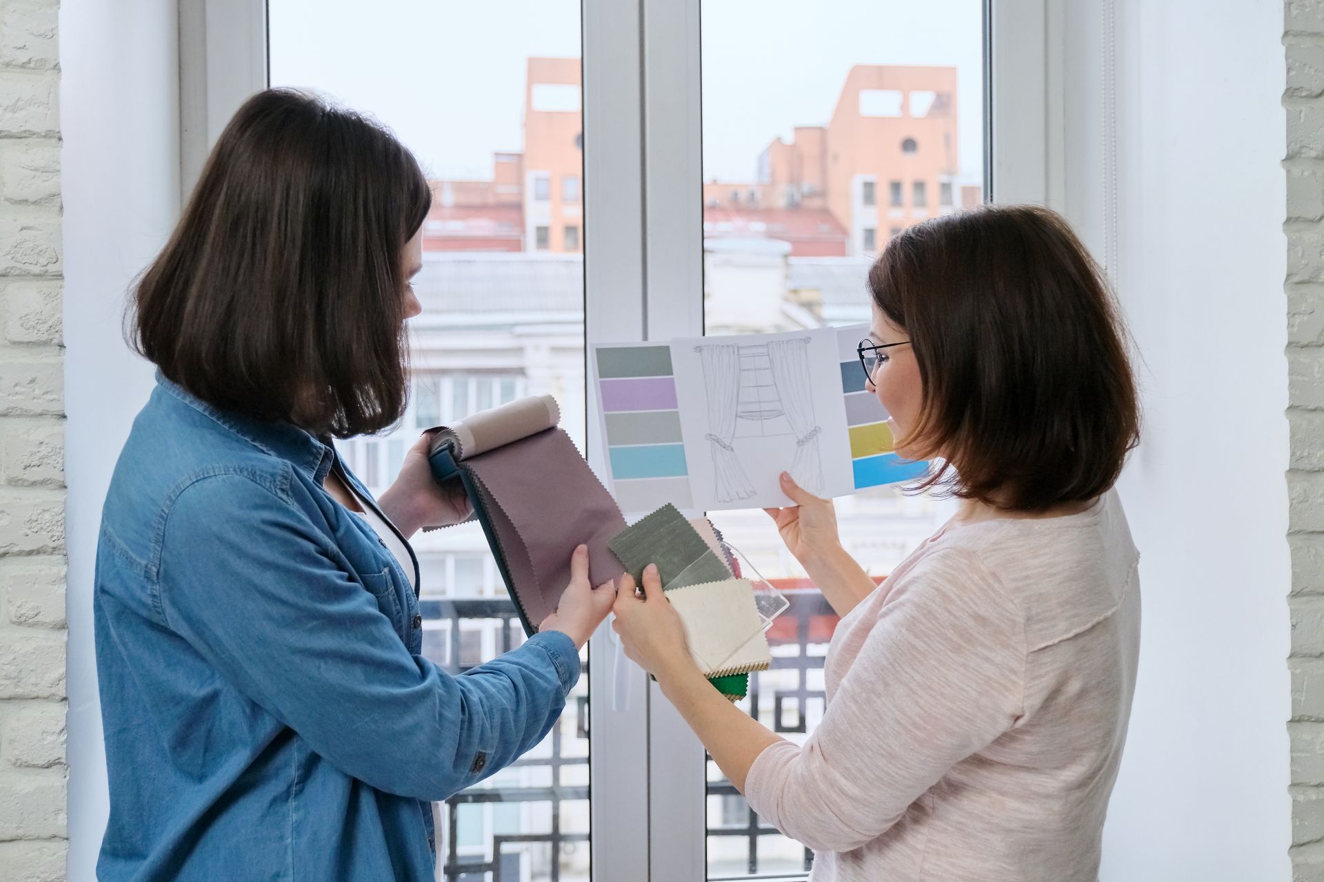 Two women are looking at fabric samples in front of a window.