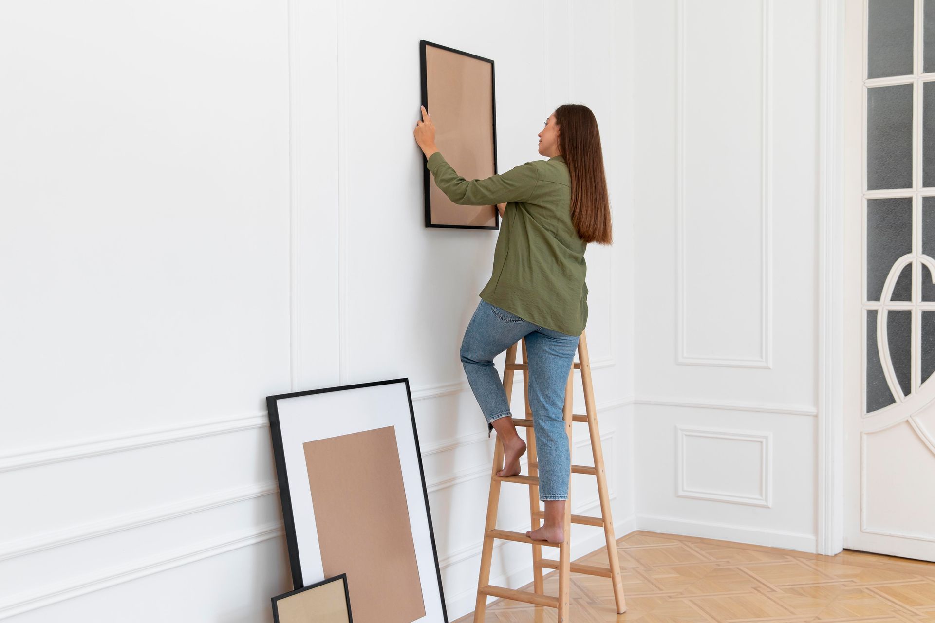 A woman is standing on a ladder hanging a picture on a wall.