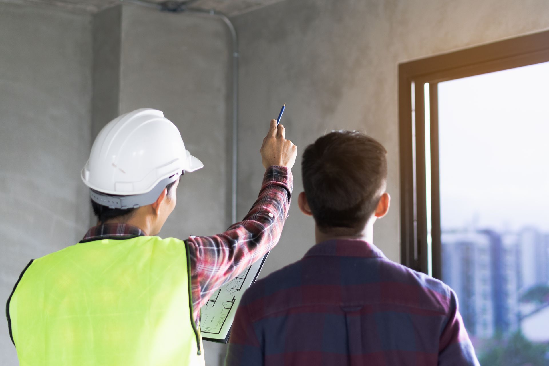A man in a hard hat is pointing at a window while another man looks on.