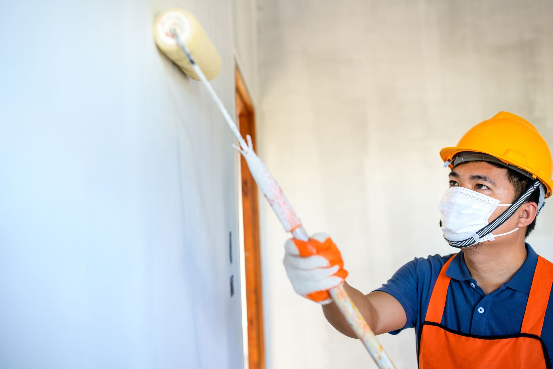 A man wearing a mask and hard hat is painting a wall with a paint roller.