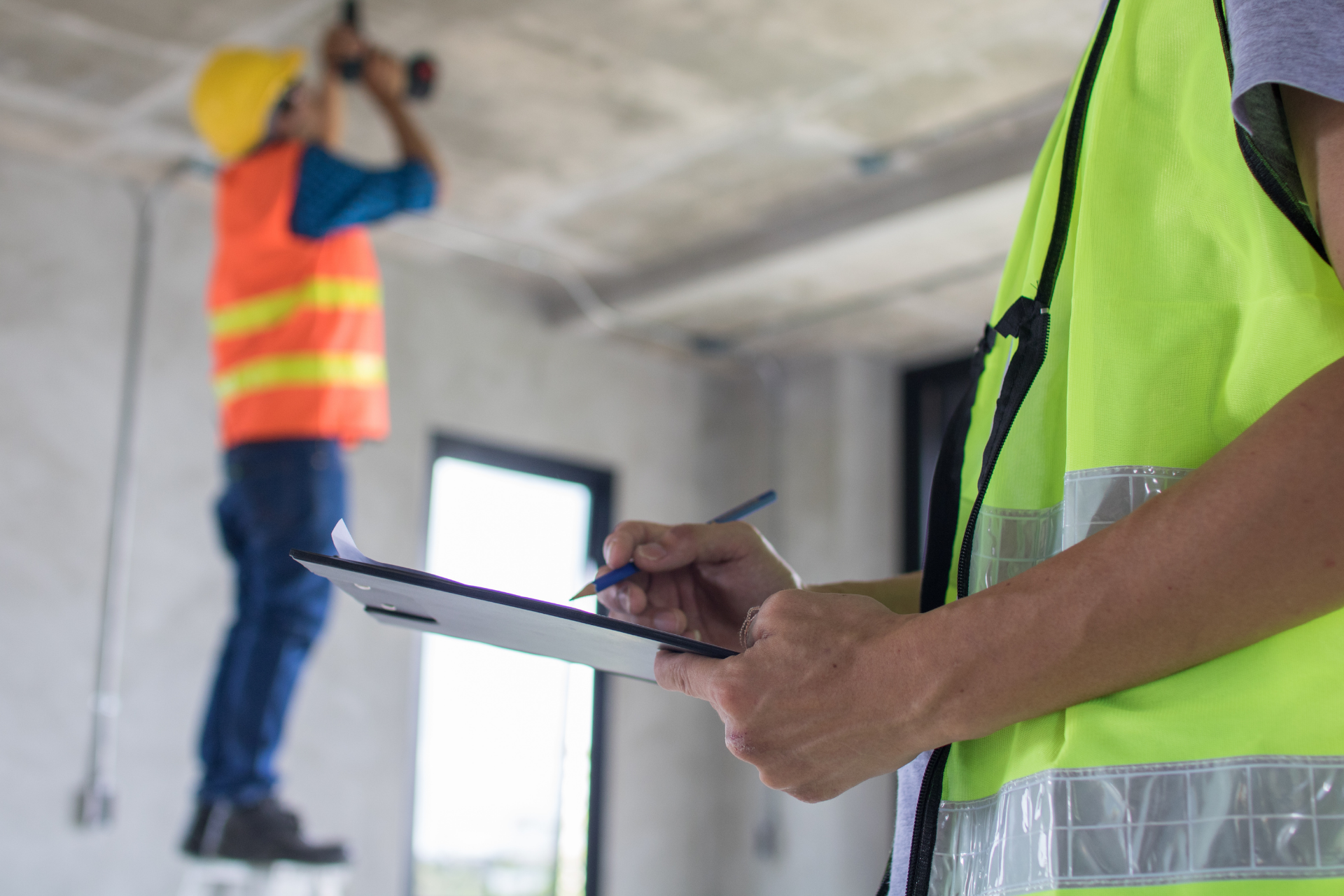 A construction worker is writing on a clipboard while another worker is working on the ceiling.