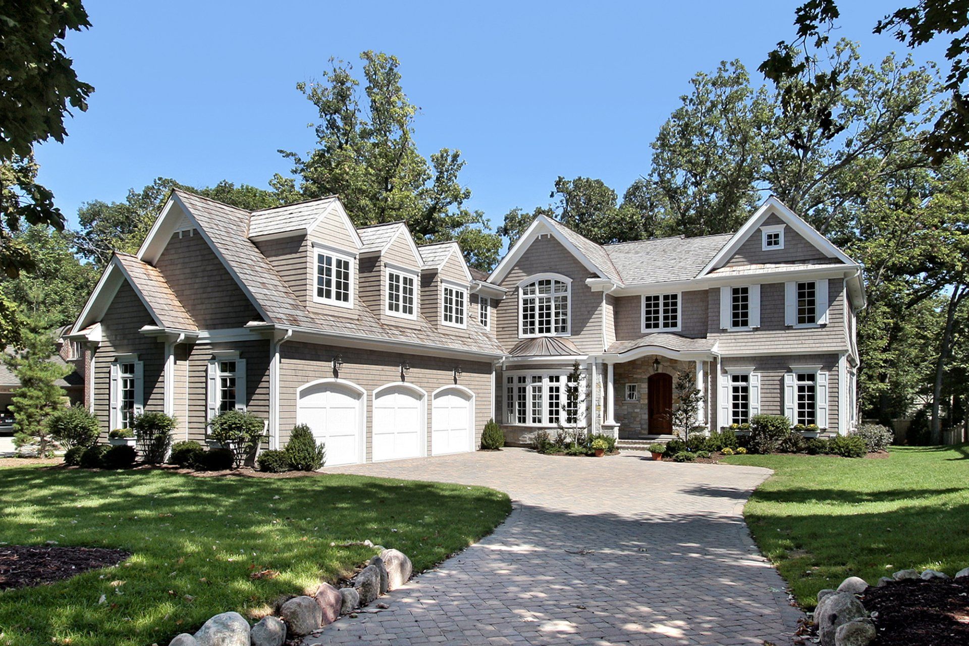 Large gray and white two-story house with a stone driveway, three-car garage, and lush green lawn under a blue sky.