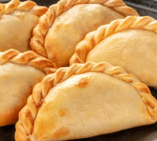 Empanadas de color marrón dorado, con bordes trenzados, dispuestas muy juntas sobre una superficie oscura.