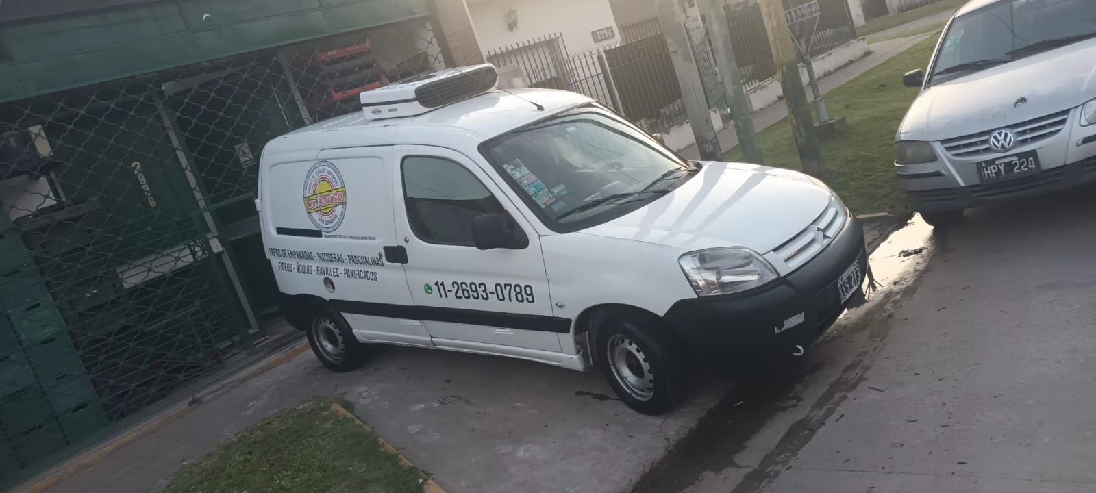 Camioneta blanca estacionada en una calle con un logo y número de teléfono en el costado, al lado de un auto plateado.