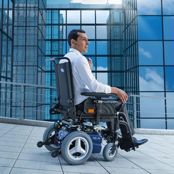 Un uomo su una sedia a rotelle elettrica sorride, osservando lo sfondo di un moderno edificio in vetro.