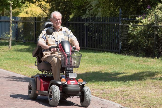 Un uomo anziano sorridente guida uno scooter elettrico rosso su un sentiero asfaltato vicino a una recinzione.