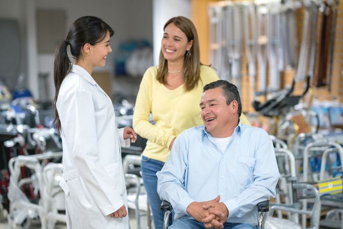 Una donna in camice da laboratorio aiuta un uomo sulla sedia a rotelle e una donna in un negozio di forniture mediche.