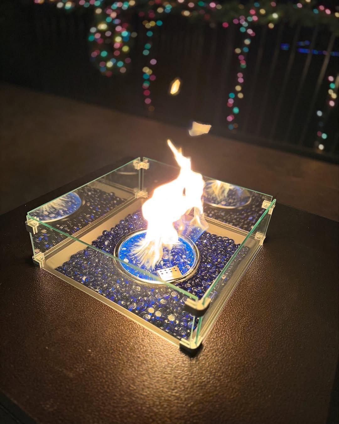 Small tabletop fire pit with blue glass beads, flames rising, and blurred lights in background.