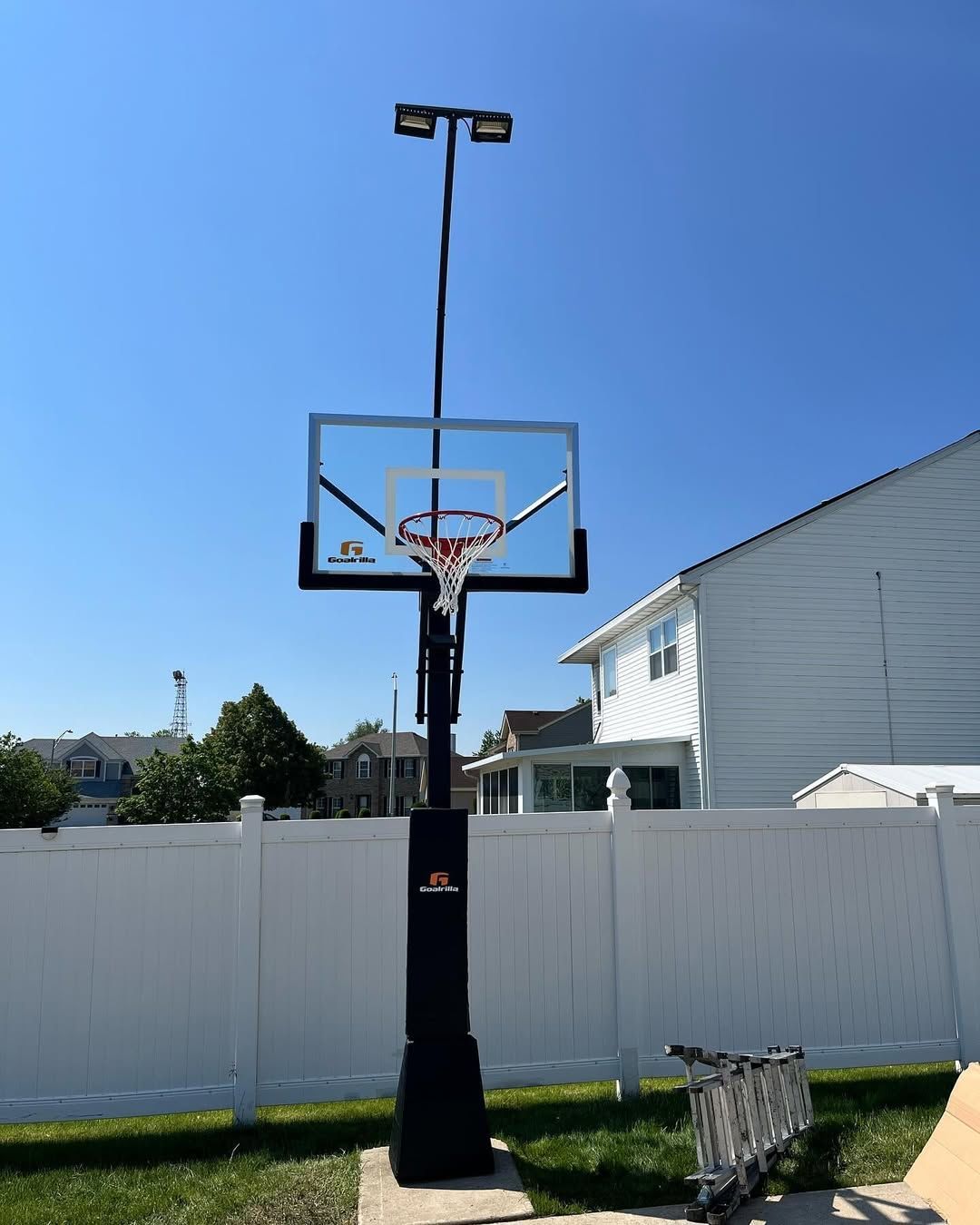 Basketball hoop with lights in a backyard, with a white fence and house in the background.