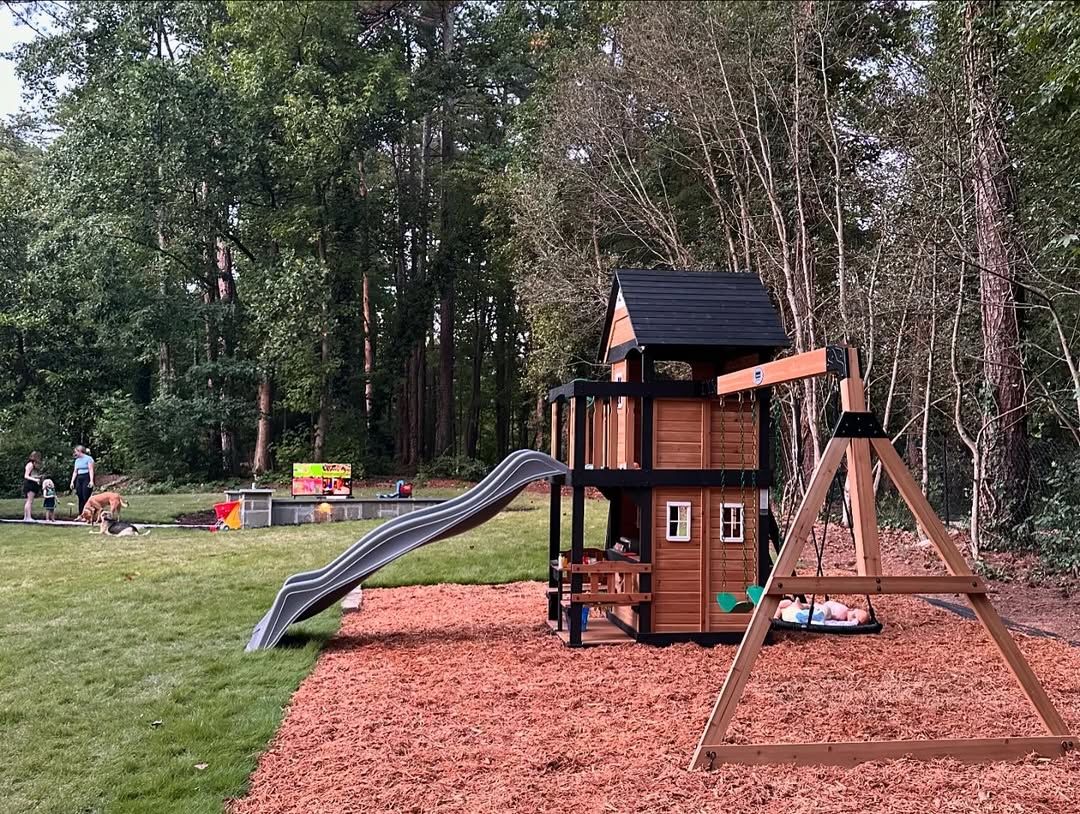 Playground with slide and swing set in a wooded area with green grass and red mulch.