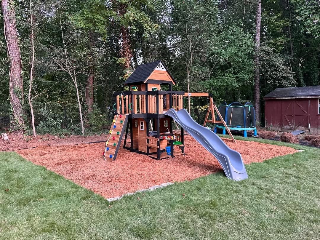 Play set in a yard with a slide, climbing wall, swing set, trampoline, and shed; surrounded by mulch and trees.