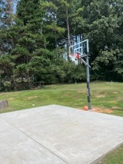 A concrete basketball court with a hoop and a ball, surrounded by grass and trees.