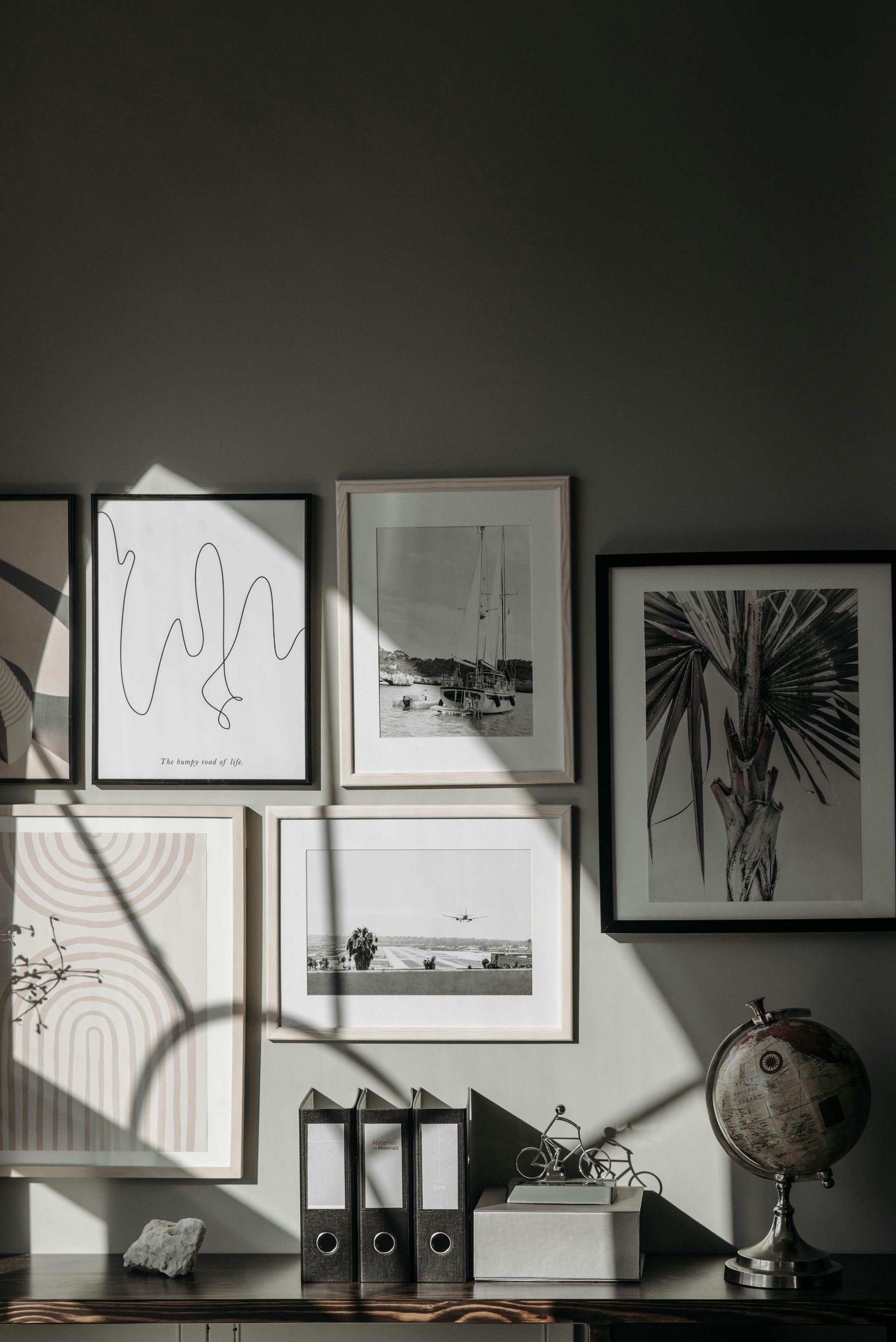 Gallery wall with framed art, desk with files, decorative globe, and sunlight shadows.