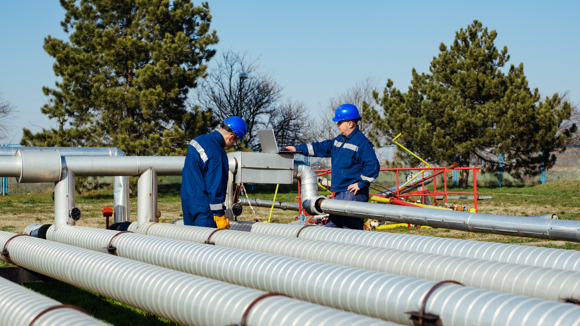 Two workers in blue coveralls inspect metal pipes outdoors.