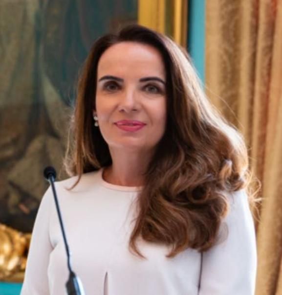 Woman with wavy brown hair, wearing a dark blazer, smiling, in front of a neutral background.