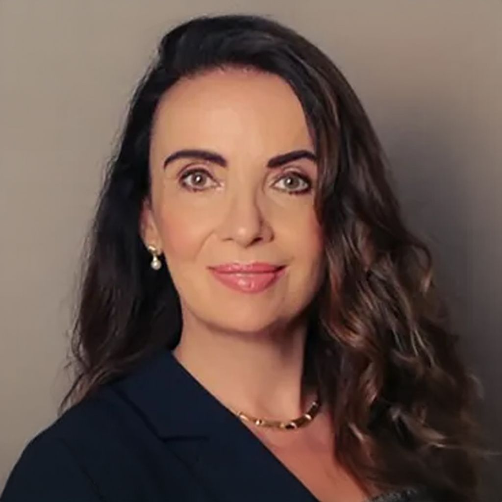 Woman with wavy brown hair, wearing a dark blazer, smiling, in front of a neutral background.