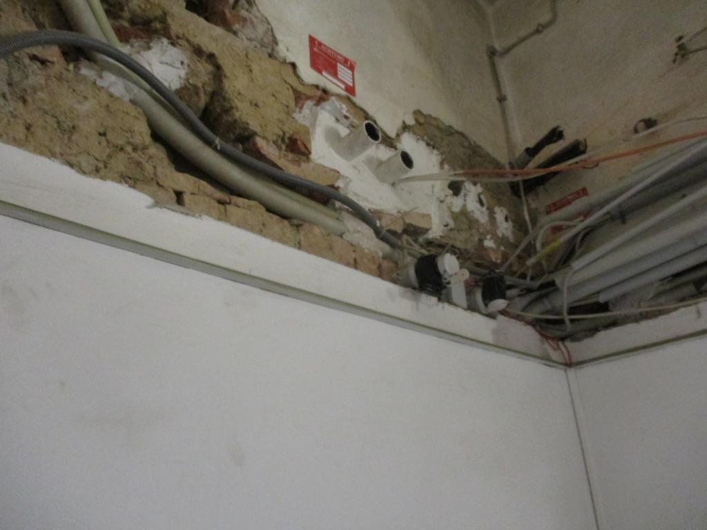 Freiliegende Kabel und Rohre in einer beschädigten Wand- und Deckenecke. Darunter eine weiße Wand.