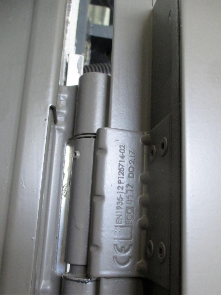 Nahaufnahme eines silbernen Türscharniers mit zylindrischem Mechanismus und einem Etikett mit Text und Zahlen.