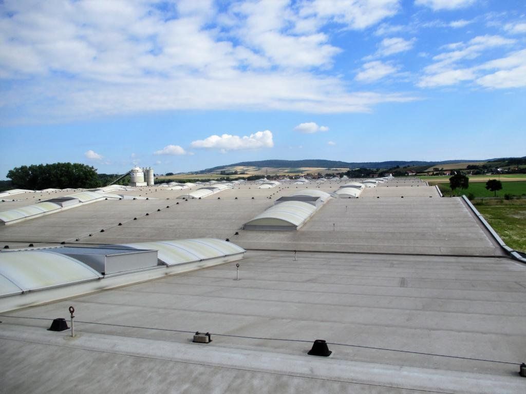 Breites, flaches Dach mit Oberlichtern unter blauem Himmel, in der Ferne ist die Landschaft zu sehen.