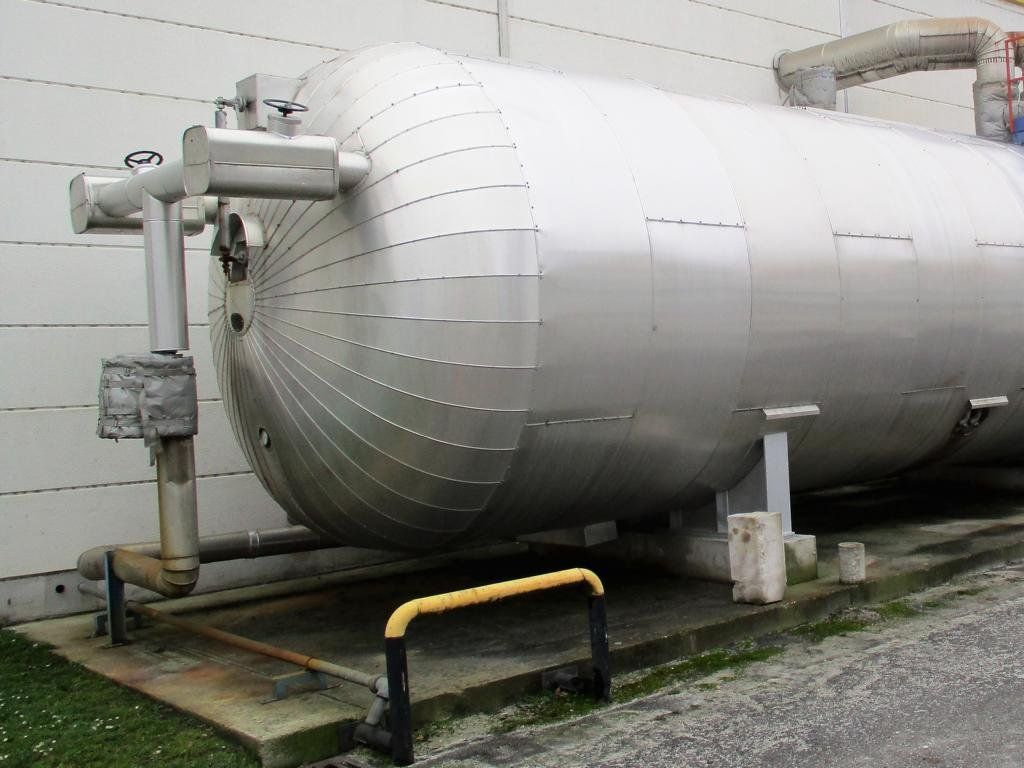 Großer, zylindrischer Metallspeichertank im Freien mit Rohren und einer gelben Sicherheitsbarriere.