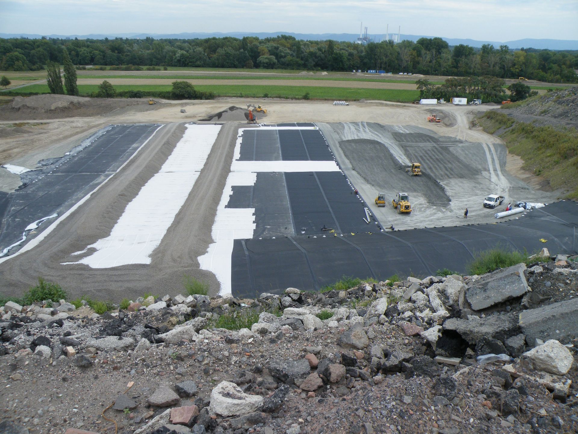 Eine Mülldeponie-Baustelle mit schwarz-weißen Abdichtungsbahnen, schweren Maschinen und Bäumen in der Ferne.