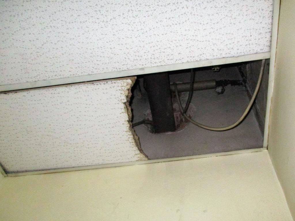 Beschädigte Deckenplatte mit einem Loch, durch das Rohre und Kabel über einer hellbeigen Wand sichtbar werden.