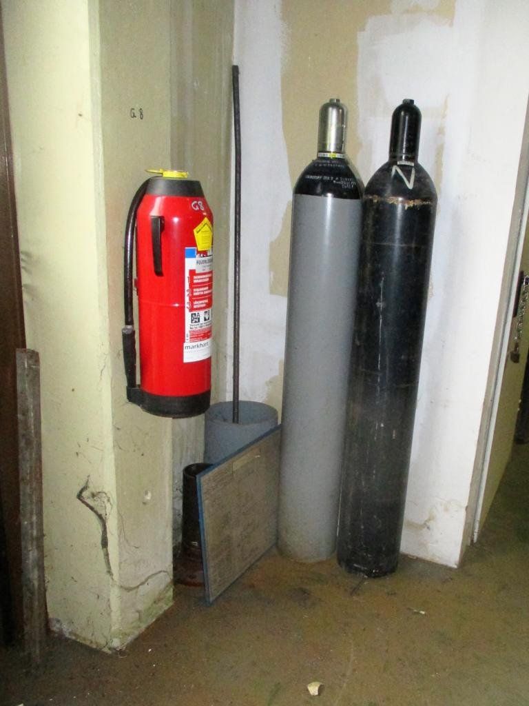 Ein roter Feuerlöscher, zwei Gasflaschen und eine Metallstange stehen in der Nähe einer Wand in einem Raum.