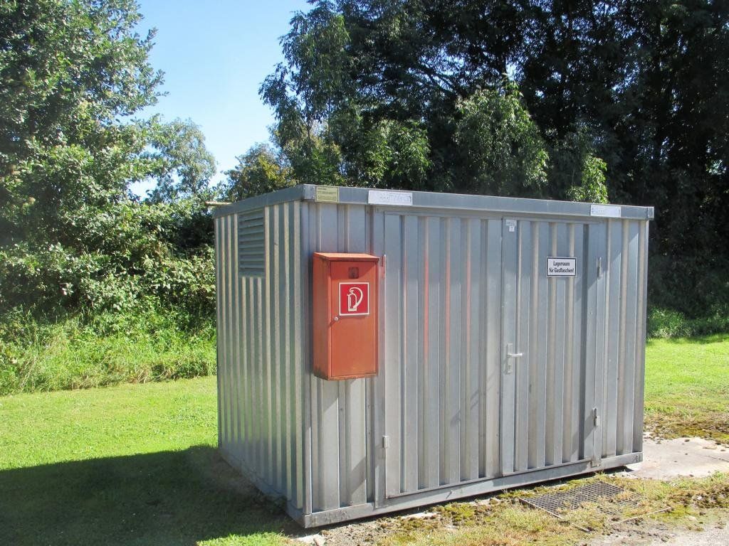 Verzinkter Metallcontainer mit Feuerlöscherkasten auf einer Rasenfläche, im Hintergrund Bäume.