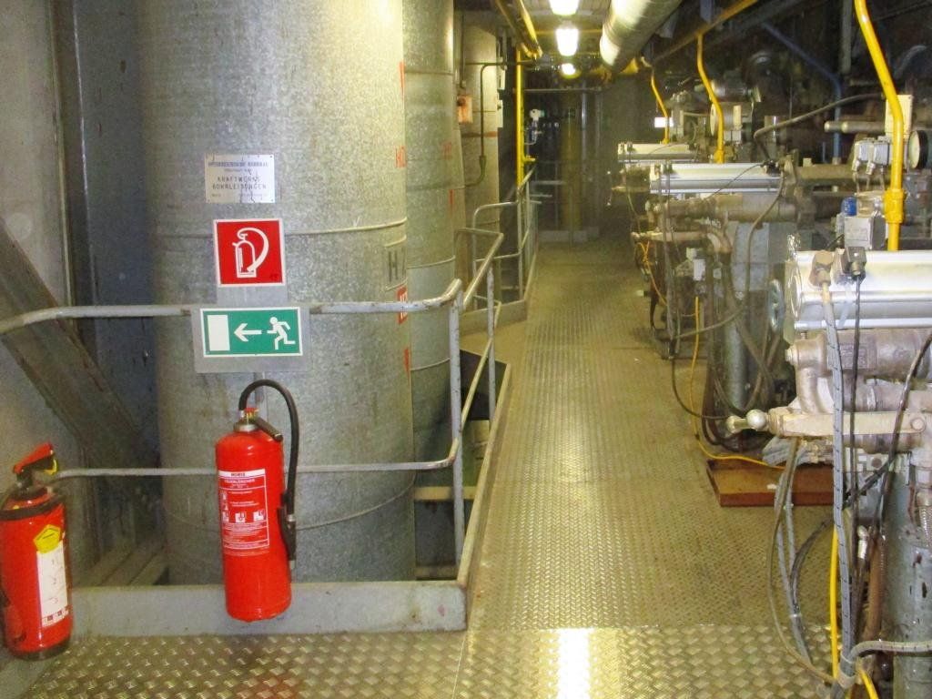Industrieinterieur mit Rohren, Maschinen, Sicherheitsschildern und Feuerlöschern in einer industriellen Umgebung.