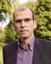 Mann mit Brille, Blazer und gestreiftem Hemd im Freien, der in die Kamera schaut.