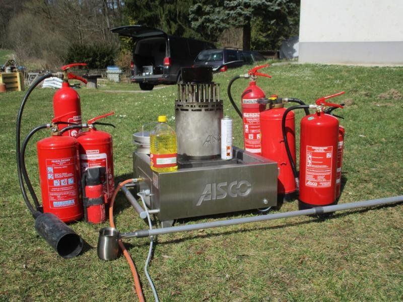 Feuerlöscher umgeben ein Metallgerät auf einer Rasenfläche; im Hintergrund ist ein schwarzer Lieferwagen zu sehen.