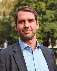 Mann im Anzug und offenem blauen Hemd, im Freien, schaut in die Kamera.