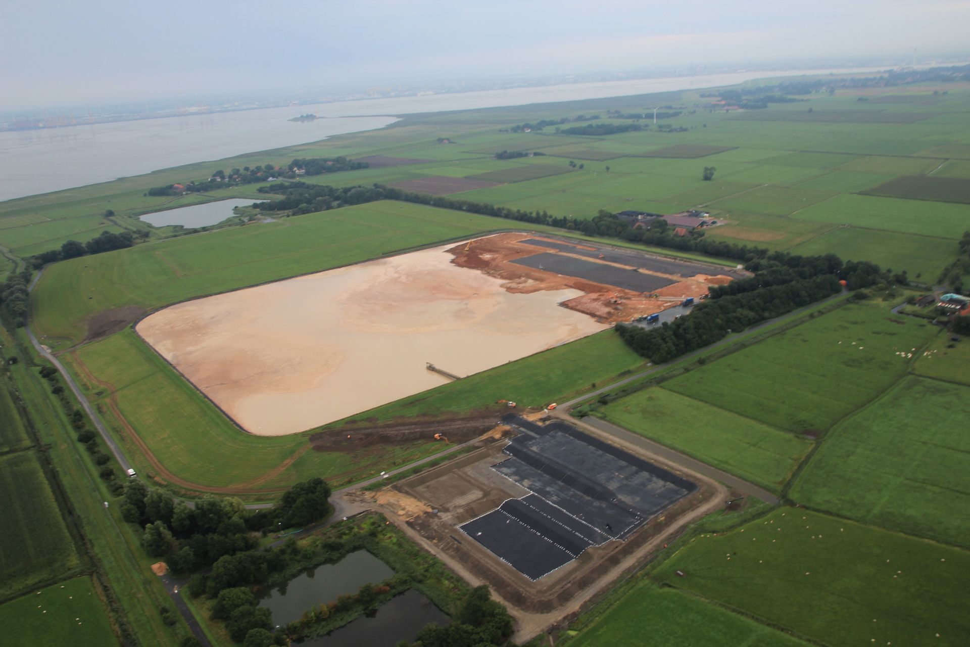 Luftbild einer braun-grünen Landschaft mit einem großen schlammigen Teich und angrenzendem Industriegebiet.