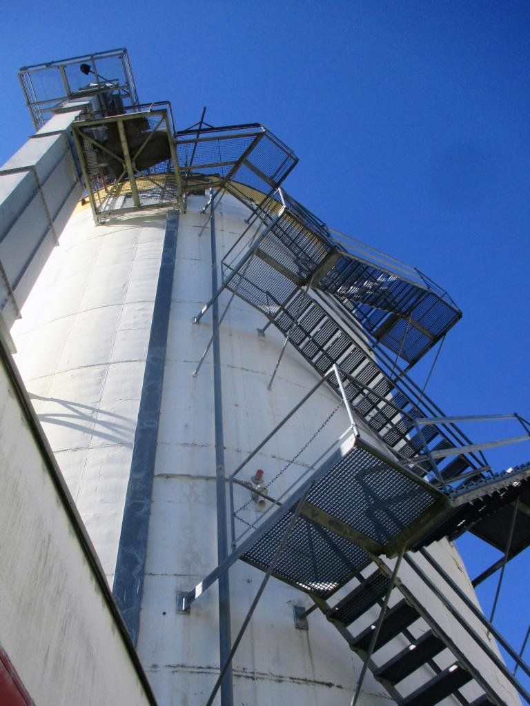 Hohes, zylindrisches Industriegebäude mit außenliegender Metalltreppe vor blauem Himmel.