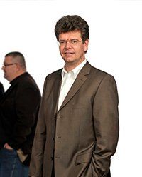 Mann in braunem Anzug, weißem Hemd und Brille, steht und lächelt. Menschen im Hintergrund.