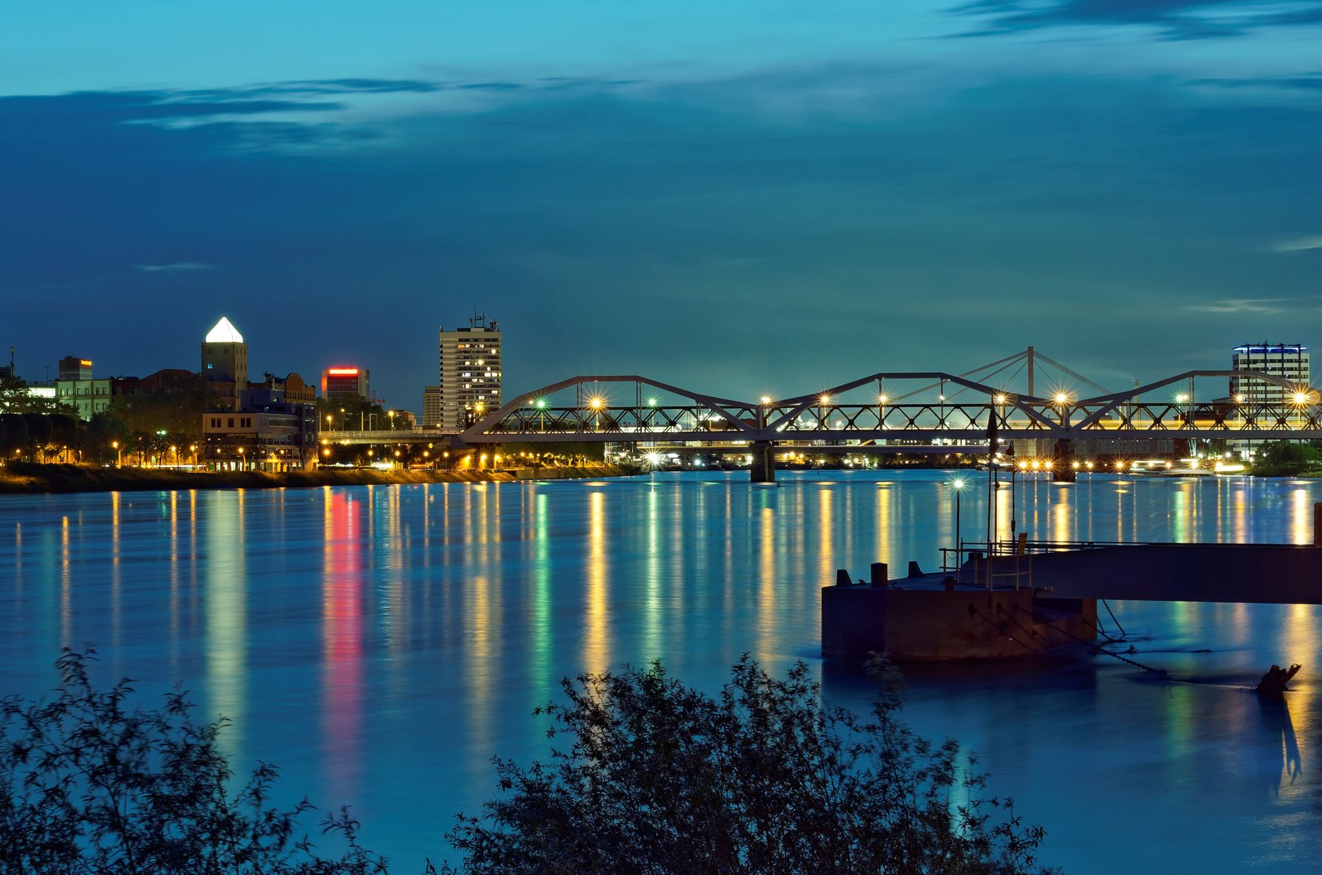 Nächtliches Stadtbild, gespiegelt im Wasser, mit Brücke und beleuchteten Gebäuden. Dunkelblauer Himmel.