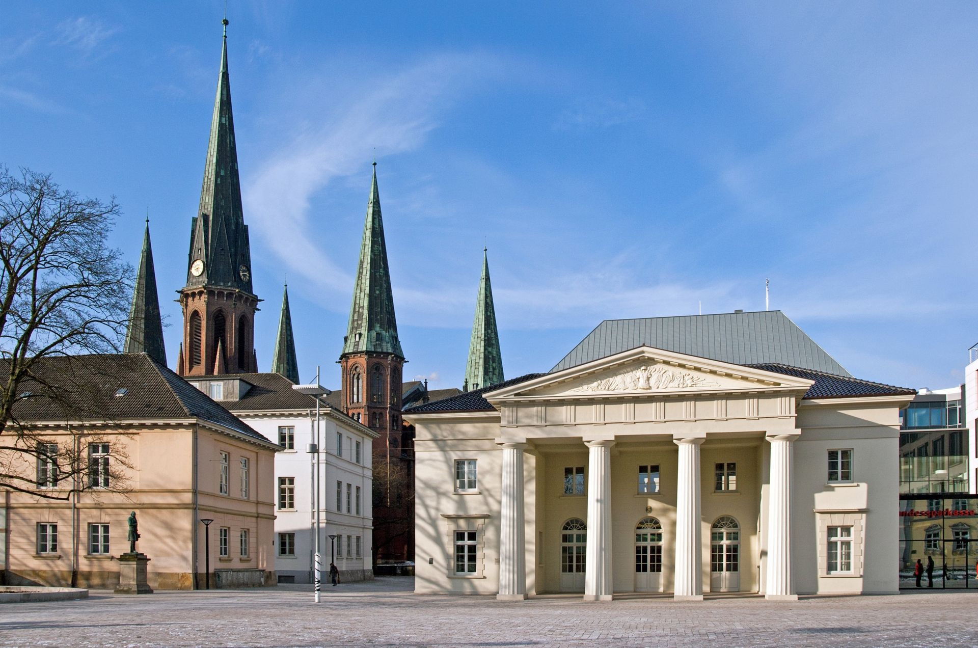 Ein neoklassizistisches Gebäude mit Säulen vor mehreren Kirchtürmen vor blauem Himmel.